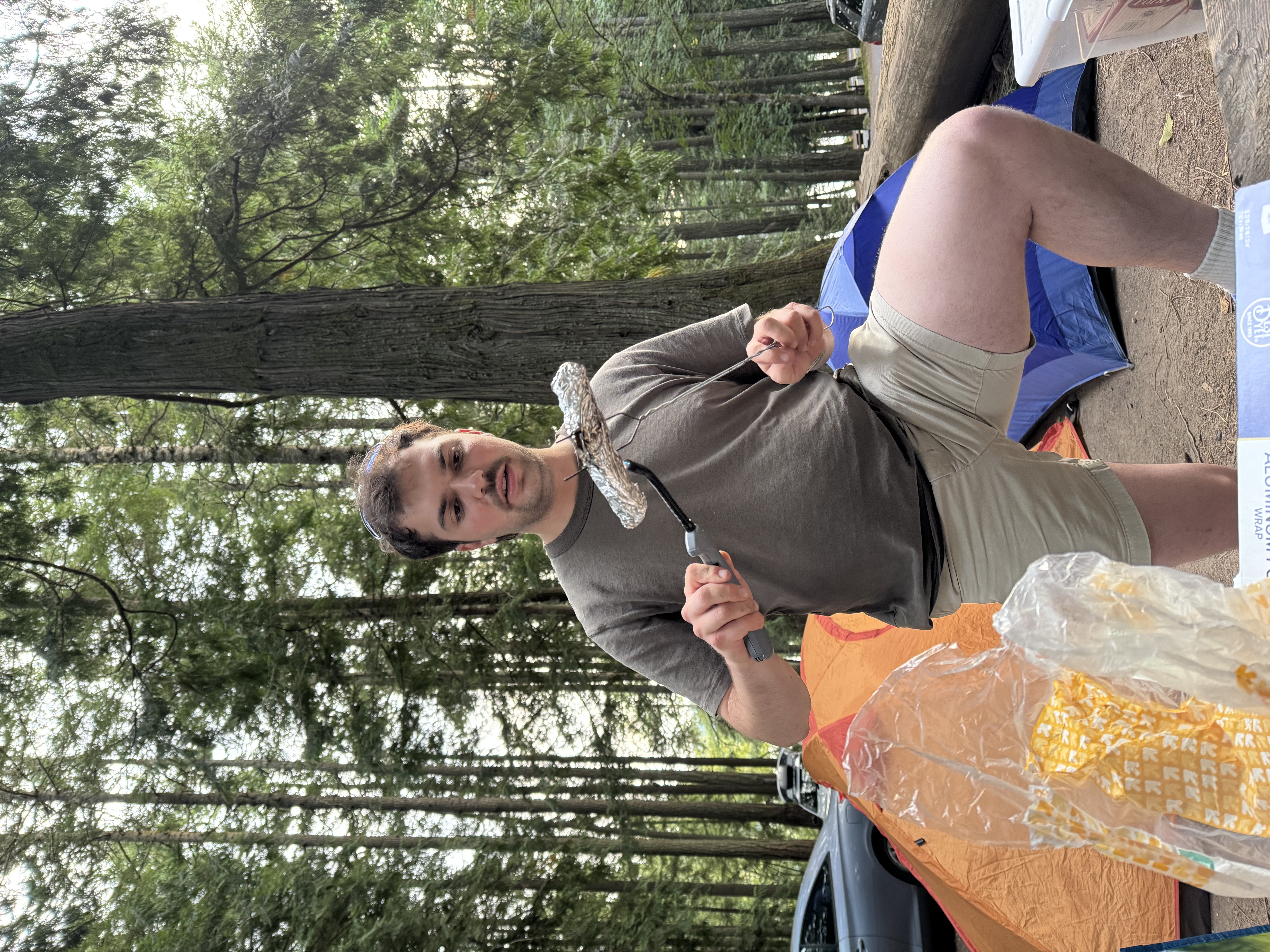 Tom Mikolyuk standing up in front of a canopy of trees and behind a picnic table, one leg up on the bench, holding a vegan hotdog wrapped in aluminum foil with a skewer in one hand and warming it up with a gas lighter held in the other hand.