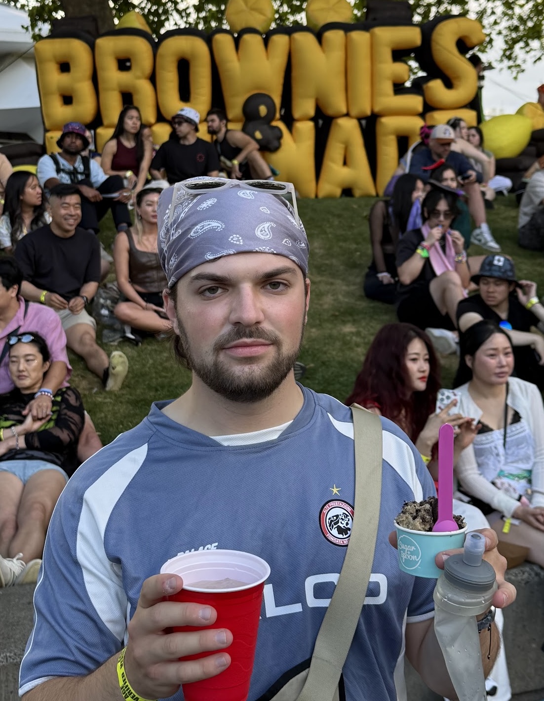 Tom Mikolyuk, wearing a gray bandana and sunglasses pushed up on his head, along with a blue and white soccer jersey, stands facing the camera at an outdoor EDM festival called 'Brownies and Lemonade'. He holds a red plastic cup in one hand and, in the other, a small cup of edible brown & black cookie dough with a purple spoon alongside a water bottle. A crowd of festival-goers sits on grass behind him, and large inflatable letters spelling 'BROWNIES & LEMONADE' (partially obscured) rise above the scene.