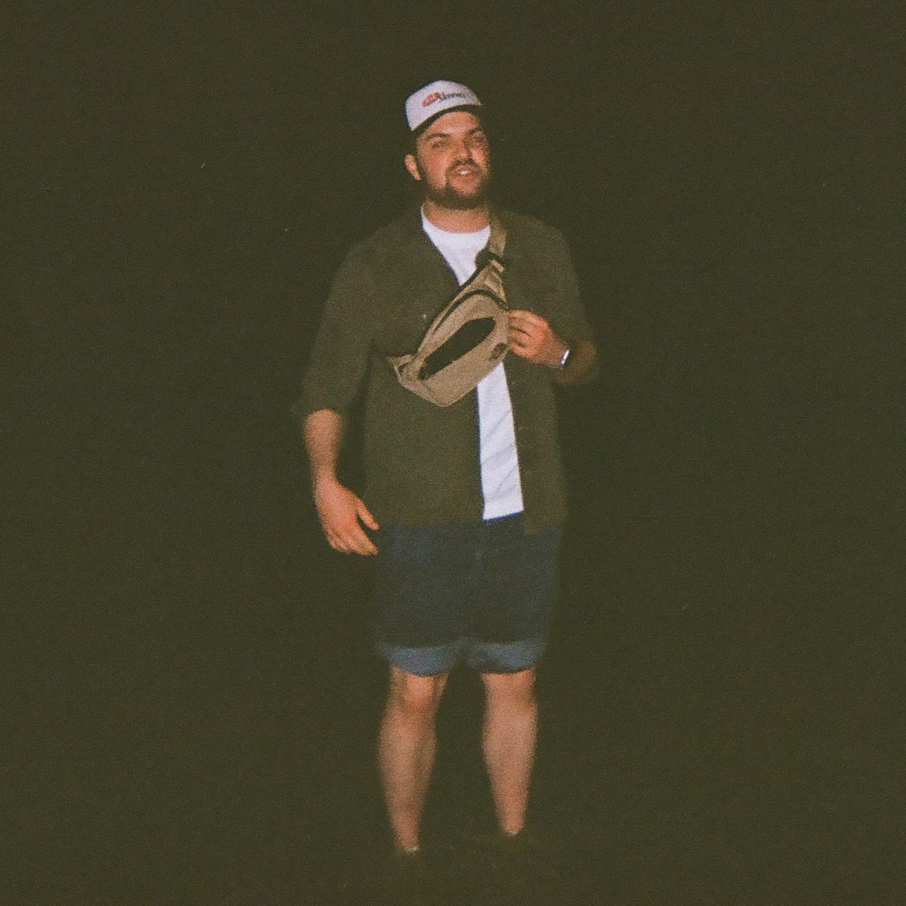 Tom Mikolyuk standing up, ankle-deep in ocean-water, in nighttimne lighting and against a dark background. He is wearing rolled-up blue jeans, e, one leg up on the bench, holding a vegan hotdog wrapped in aluminum foil with a skewer in one hand and warming it up with a gas lighter held in the other hand.