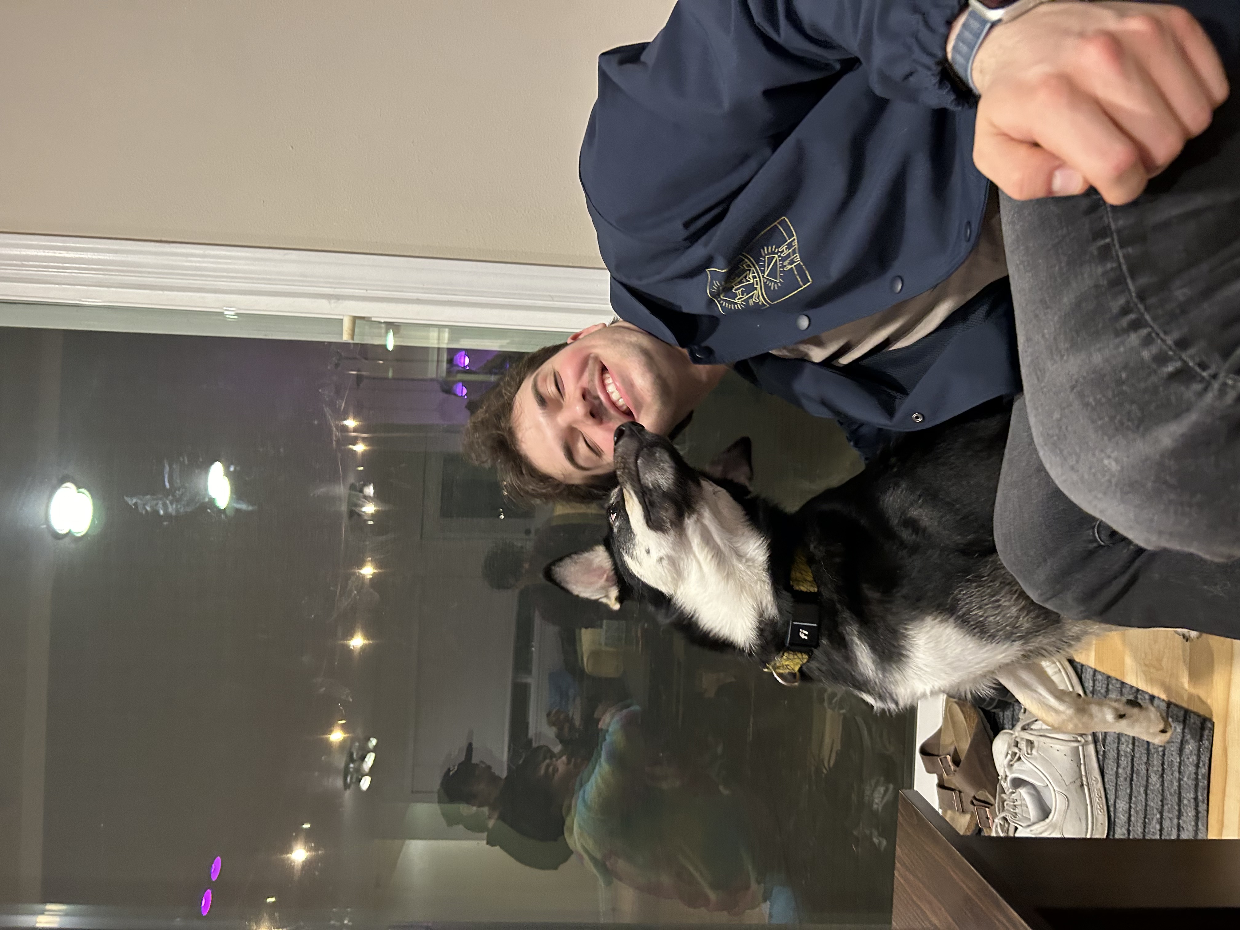 Tom Mikolyuk sitting on a couch, his face being licked by a large black dog sitting on the floor next to the couch.